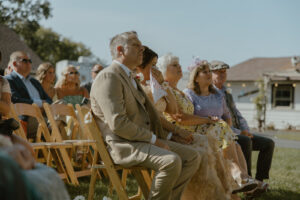 Side angle view of outdoor backyard wedding ceremony guests seated on wooden folding chairs on lawn