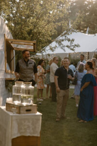Wedding guests laughing at vintage camper bar with server in patterned vest and mason jar drink dispensers at golden hour