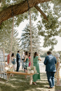 Wedding cocktail hour guests mingling under oak tree with rope swing bench floral arrangements and welcome sign