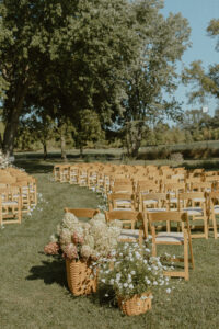 Outdoor backyard wedding ceremony setup with rows of wooden chairs on lawn wildflower baskets lining the aisle and oak trees
