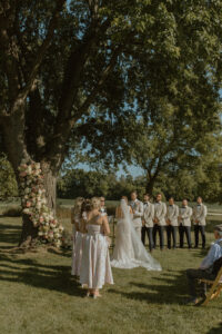 Full wedding party portrait standing together under large oak trees at backyard wedding