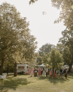 Wide golden hour shot of backyard wedding cocktail hour guests mingling around vintage white camper bar with bunting under oak trees