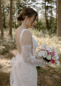Three quarter back portrait of bride in white lace open back wedding dress holding pink bouquet in pine forest