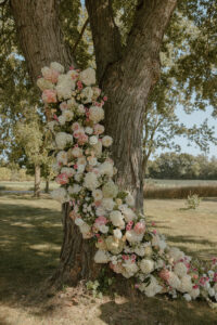 White cream and pink hydrangeas and dahlias cascading up the split trunk of a large oak tree wedding floral installation
