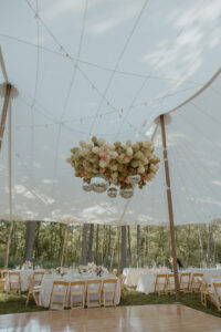 Wide interior view of backyard wedding sailcloth tent with floral disco ball installation round tables set and forest beyond