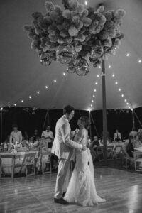 Black and white photo of bride and groom first dance under string lights at night with guests seated at tables